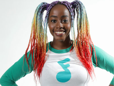 Aiza, a Black woman with her hair in colourful braids, smiles at the camera. She is wearing a white t-shirt with teal arms and a teal Jeremy and Jazzy logo.